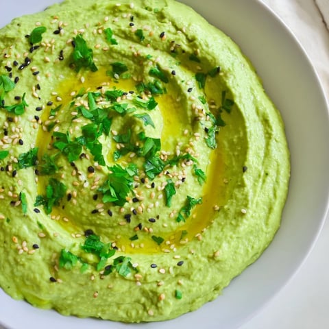 A bowl of vibrant green Edamame Hummus garnished with sesame seeds and parsley, served with pita chips and fresh vegetables.