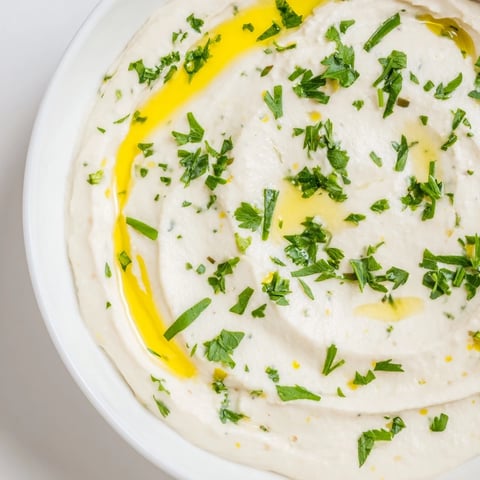A bowl of creamy White Bean Dip drizzled with olive oil and parsley, served with pita and colorful vegetable sticks for dipping.  