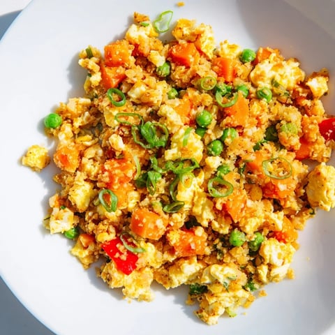 A close-up of cauliflower fried rice with vibrant orange carrots, green peas, and scrambled eggs piled high on a white plate.  