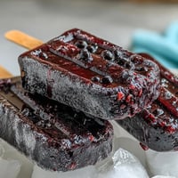 Homemade Black Currant Popsicles with vibrant purple hue and fresh fruit chunks, resting on a chilled plate for a refreshing vegan summer dessert.